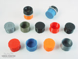 All colour variations of forster uk's CRF mount outer bayonet deep rear lens caps are being displayed on a plain white background. The caps are being shown in black, orange, red, mid blue, silver and survival green, all which are available for purchase on simonforsterphotographic.co.uk.