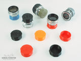 All colour variations of forster uk's CRF and Nikon S mount inner bayonet rear lens caps are being displayed on a plain white background. The caps are being shown in black, orange, red, mid blue, silver and survival green, all which are available for purchase on simonforsterphotographic.co.uk.