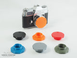 An orange DKL Schneider body cap designed to fit kodak retina reflex cameras has been fitted onto its intended camera. There more the same caps being displayed in different colours, such as black, red, mid blue, silver and survival green.