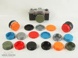 A range of rear lens caps for HI topcor and UV topcor lenses, there are also body caps which have been made to fit topcon cameras. Each type of cap has multiple of the same caps just with different colours. The colours they are all in is black, orange, red, mid blue, silver and survival green. The caps, camera and two lenses are all being displayed on a white background.